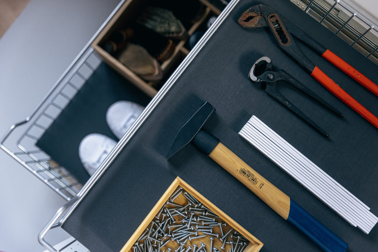 Nahaufnahme einer Werkbank mit Hammer, Zange, Metalllineal und Nagelbox auf der Antirutschmatte 'Wabe' von stauraum-shop. Im Hintergrund sind teilweise ein Regal und weiße Schuhe zu sehen.